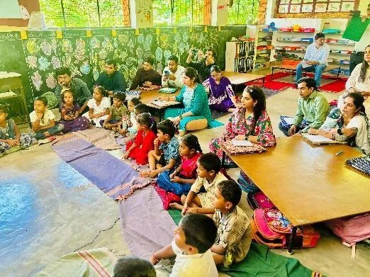 कक्षा में फर्श और नीची मेजों पर समूह में बैठे बच्चे और शिक्षक